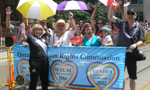 Des membres du personnel de la CODP défilent au défilé de la fierté gaie de 2012 avec la commissaire en chef Barbara Hall et la députée provinciale Cherie DiNovo. Le groupe brandit une bannière qui indique Code des droits de la personne 1962, orientation sexuelle 1986 et identité sexuelle et expression de l'identité sexuelle 2012.