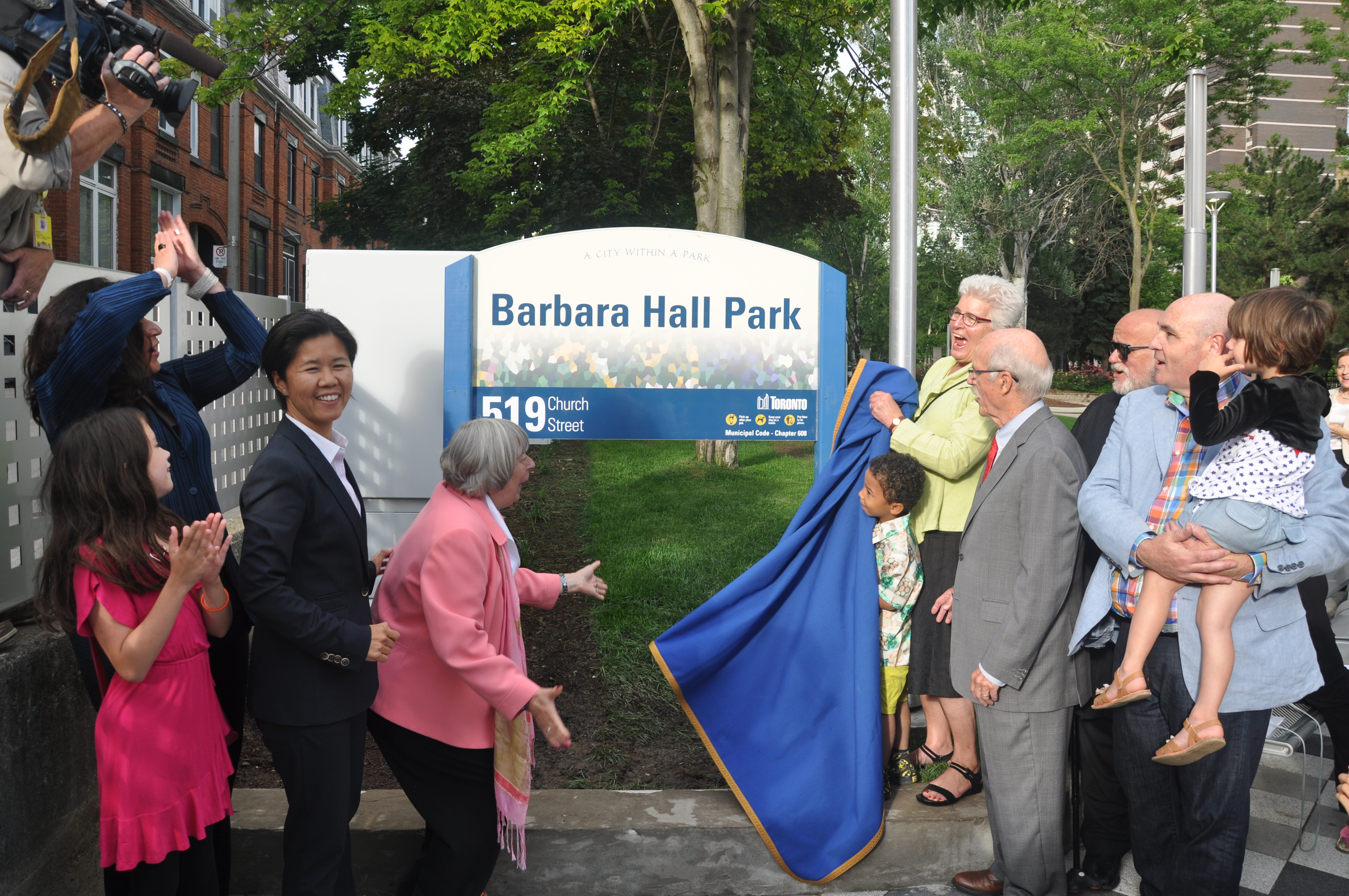 Chief Commissioner Barbara Hall unveils the new sign at Barbara Hall Park. Her Husband Max and City Counsellor Kristen Wong Tam stand in the small crowd nearby.