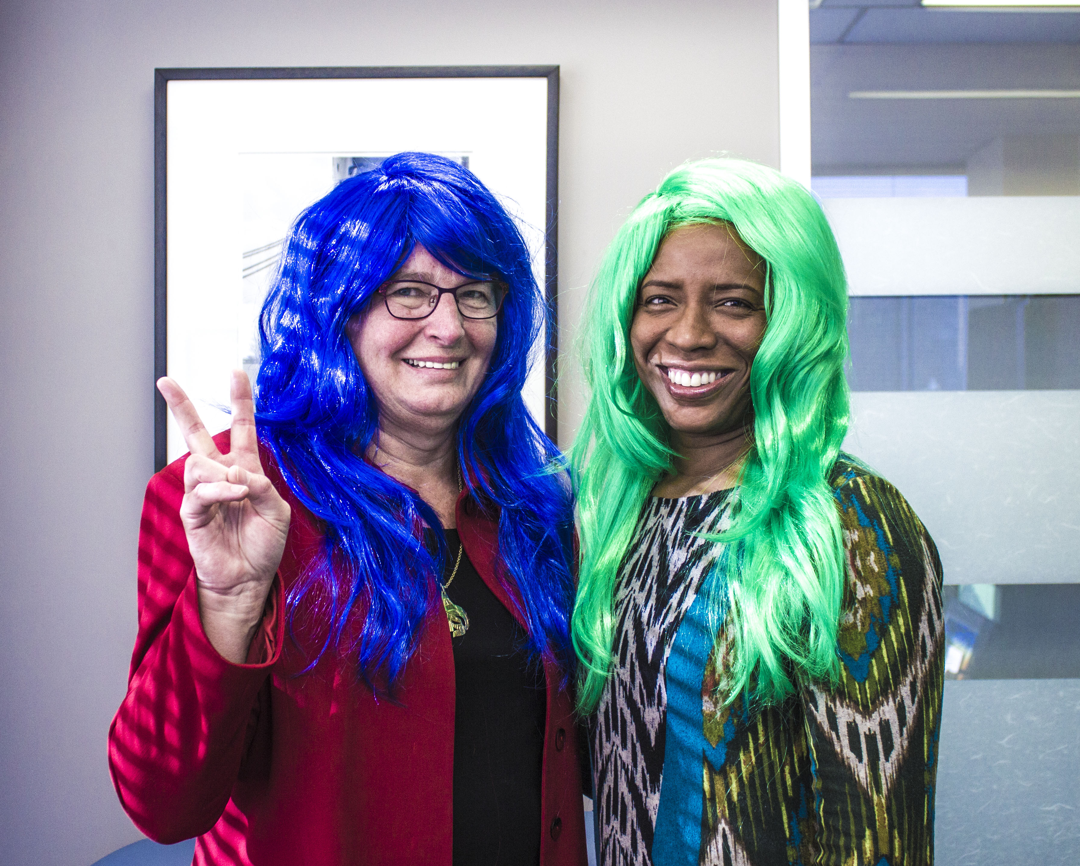 Chief Commissioner Barbara Hall stands with Executive Director Diane Carter. Diane wears a long bright green wig. Barbara wears a long, bright blue wig and holds up her peace fingers.