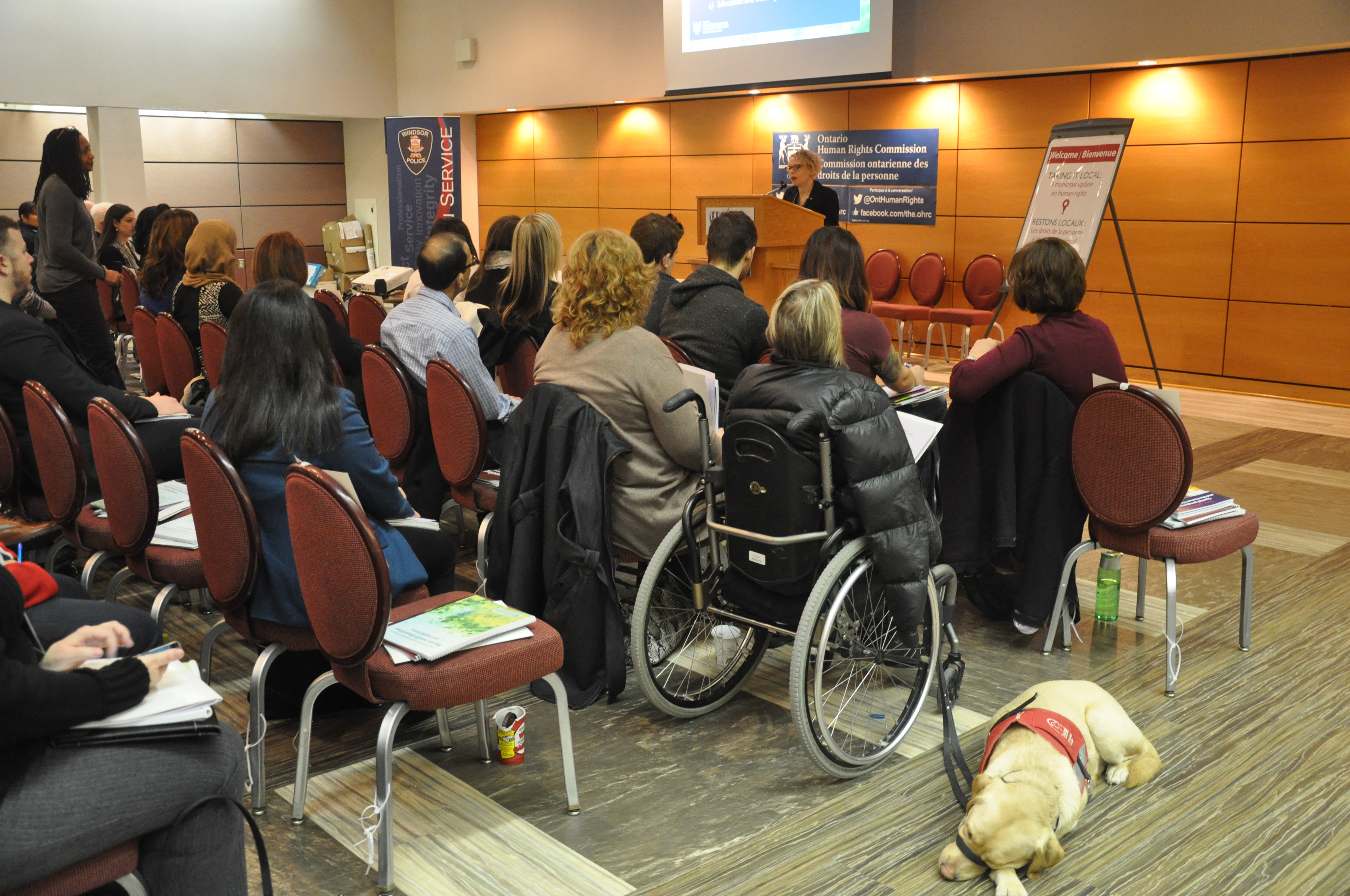 L’analyste principale des politiques, Cherie Robertson, s’adresse de la tribune à un groupe de participants. À l’avant-plan se trouvent une femme en fauteuil roulant et son chien d’assistance. 